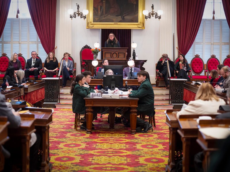 A legislative chamber with officials seated at desks, a speaker at the podium, and others seated behind; red carpets and drapes decorate the room.