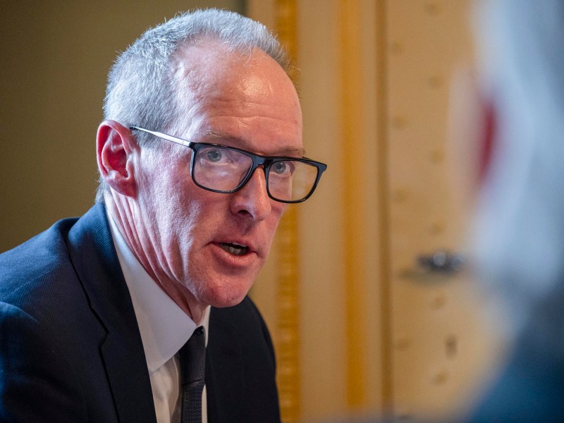 A man in a suit and glasses speaks during a meeting, with another person visible in the foreground.