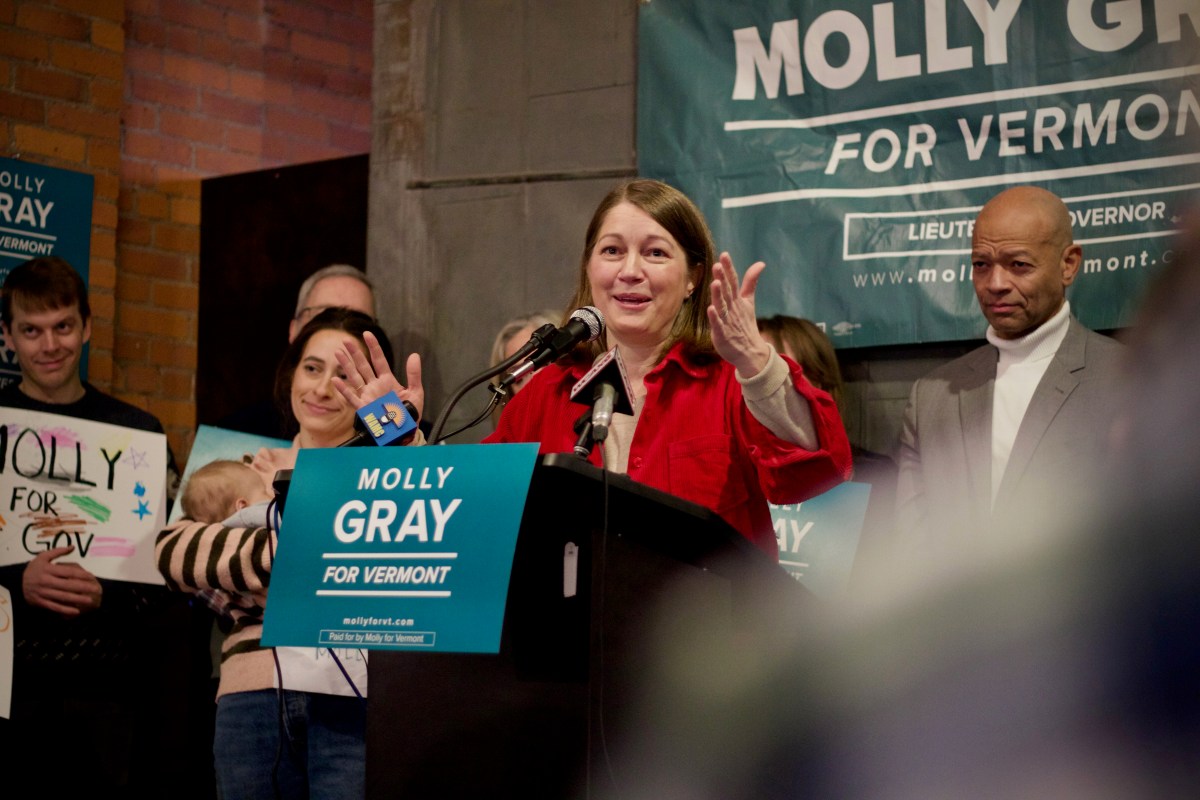 A woman speaks at a podium with "Molly Gray for Vermont" signs, surrounded by supporters at an indoor campaign event.
