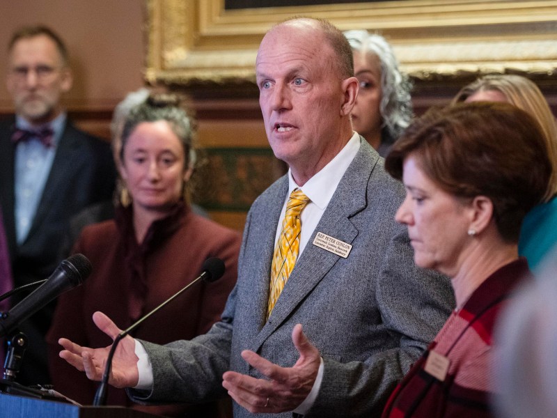 A man in a suit speaks at a podium with microphones, surrounded by several people in formal attire in an indoor setting.