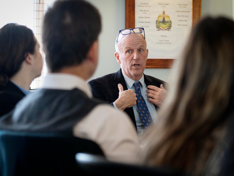 A man in a suit and blue tie speaks while gesturing with his hands during a meeting, with three other people partially visible in the foreground.