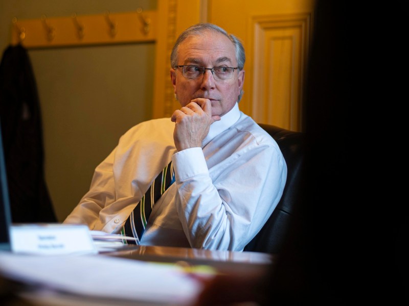 A man in a white shirt and striped tie sits at a desk, resting his chin on his hand, with papers and a nameplate in front of him.