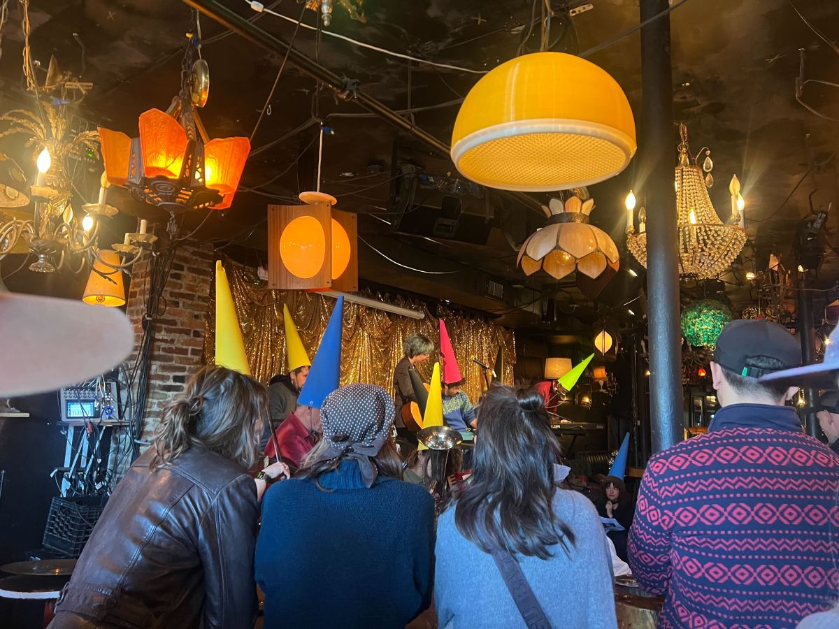 A group of people wearing colorful cone hats sit in a dimly lit room decorated with various hanging lamps and a gold tinsel backdrop, watching a stage performance.
