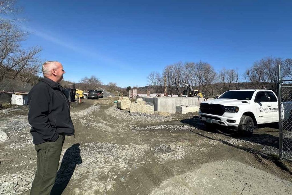 A man stands on a gravel construction site, looking at equipment and materials, with a white pickup truck parked nearby under a clear blue sky.