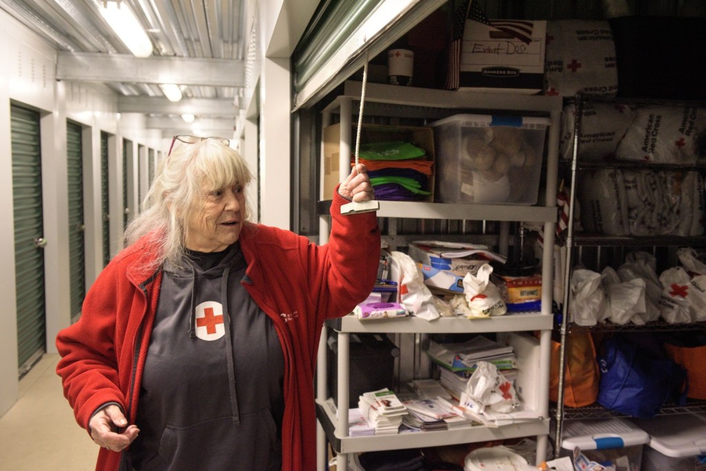 An older woman in a Red Cross jacket opens a storage unit filled with supplies and organized bins in a hallway lined with metal doors.