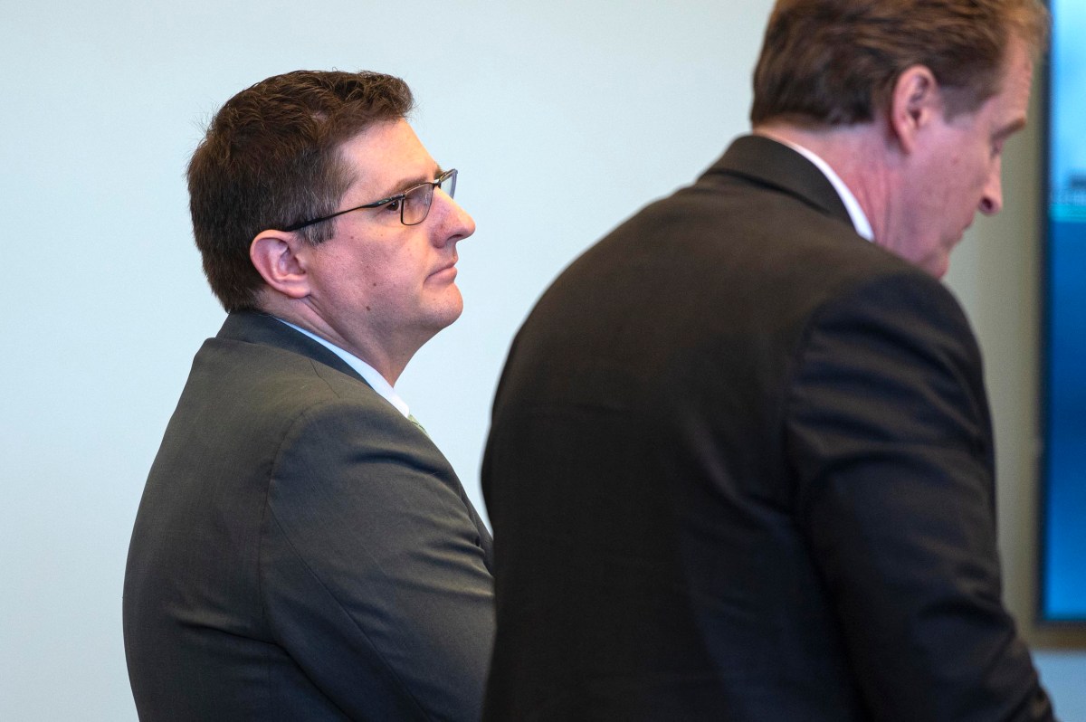 Two men in suits stand indoors, one facing forward in profile and the other turned away, both appearing attentive in a professional setting.