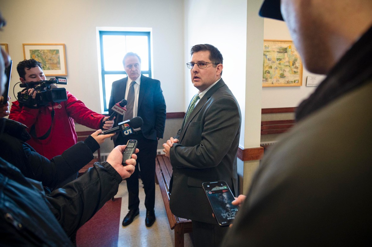 A man in a suit speaks to reporters holding microphones and recording devices in a hallway, with another man standing behind him.