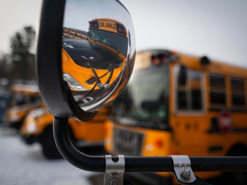 A close-up of a school bus’s side mirror reflecting the front of the bus, with other yellow school buses parked in the background.