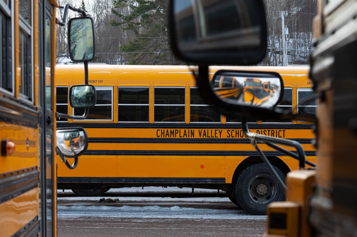 A yellow school bus labeled "Champlain Valley School District" is parked on a winter day, viewed between two other buses with mirrors visible in the foreground.
