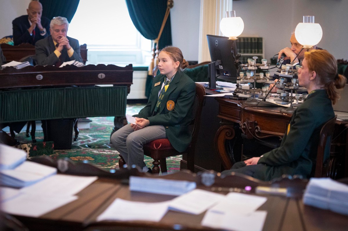 A young person in a green blazer speaks while seated in a formal meeting room, surrounded by adults and desks with papers and lamps.