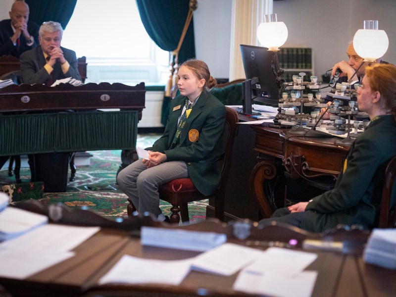A young person in a green blazer speaks while seated in a formal meeting room, surrounded by adults and desks with papers and lamps.