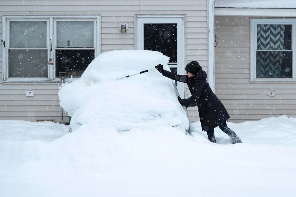 A person in winter clothing clears deep snow from a car parked in front of a beige building during snowfall.
