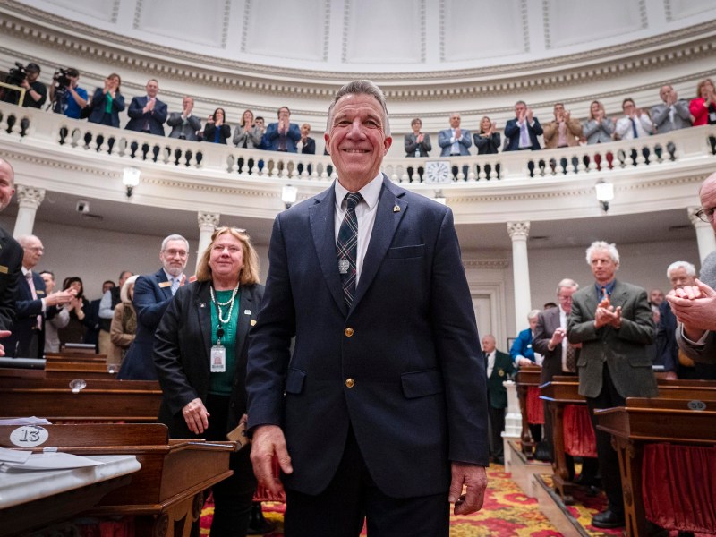 A man in a suit stands smiling in a government chamber while people around him applaud and others watch from a balcony above.