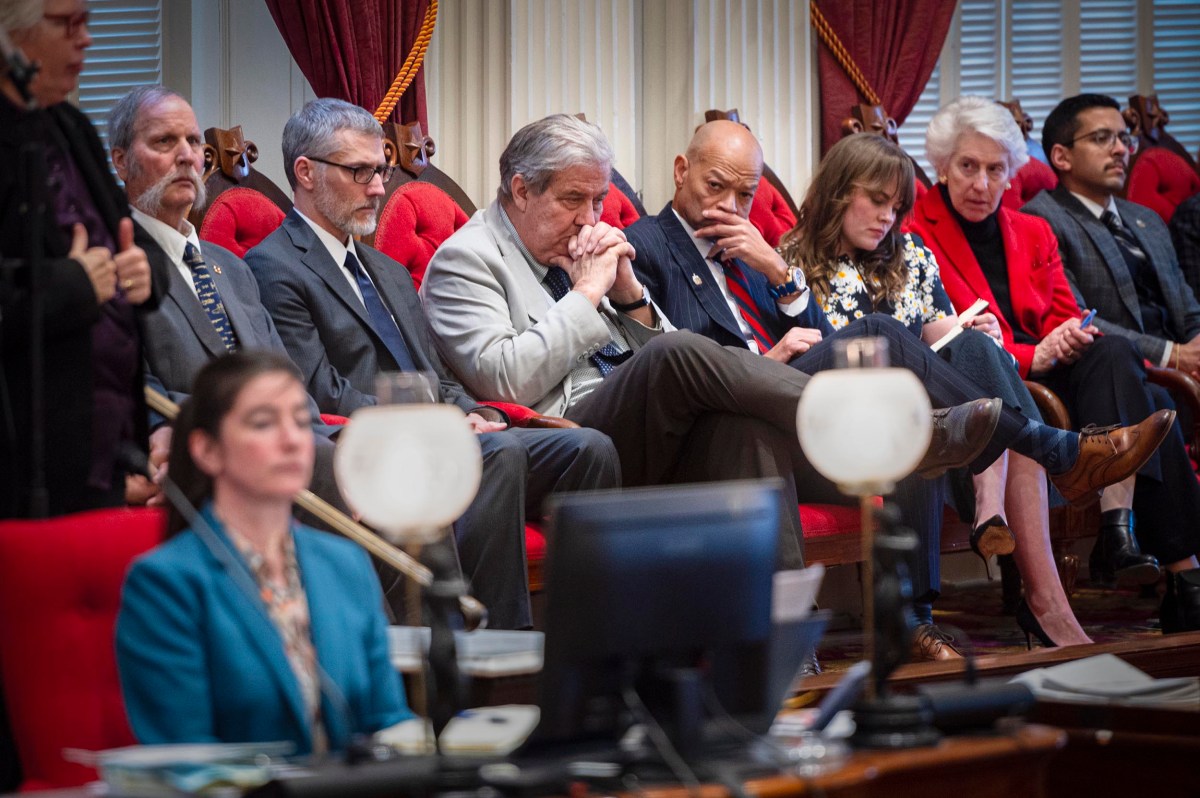 A group of people sit in red chairs in a formal setting, some looking thoughtful or serious, with documents and computers visible in the foreground.
