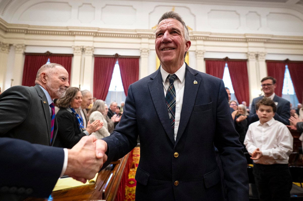 A man in a suit shakes hands with someone in a formal room as people stand and applaud in the background.