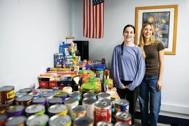 Two women stand next to a table filled with canned and packaged food items in a room with an American flag and framed artwork on the wall.