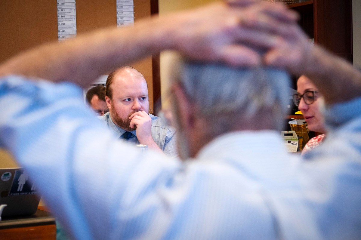 A man sits with his hand on his chin, appearing thoughtful, while another person in the foreground holds their hands on their head. Two other people are partially visible.