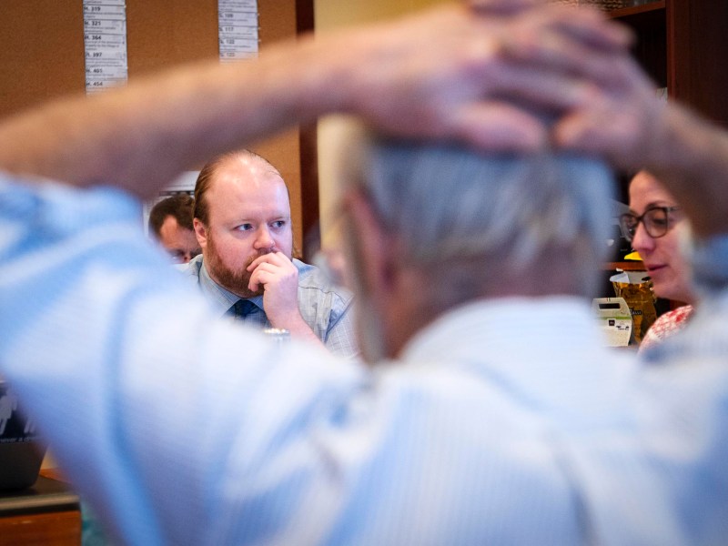 A man sits with his hand on his chin, appearing thoughtful, while another person in the foreground holds their hands on their head. Two other people are partially visible.