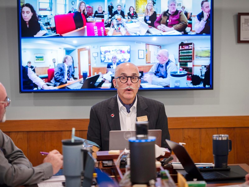 A man in a suit speaks at a conference table with laptops, while two video calls with multiple participants are displayed on a large screen behind him.