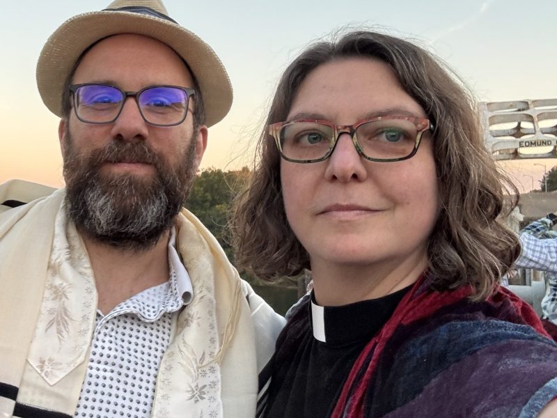 Two people wearing glasses pose for a photo outdoors at sunset; one wears a hat and scarf, the other wears a clerical collar. People and a structure are visible in the background.