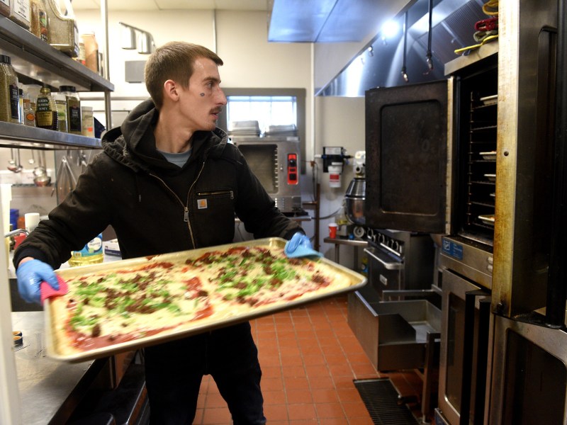 A man wearing gloves places a large rectangular pizza on a baking tray into an industrial oven in a commercial kitchen.