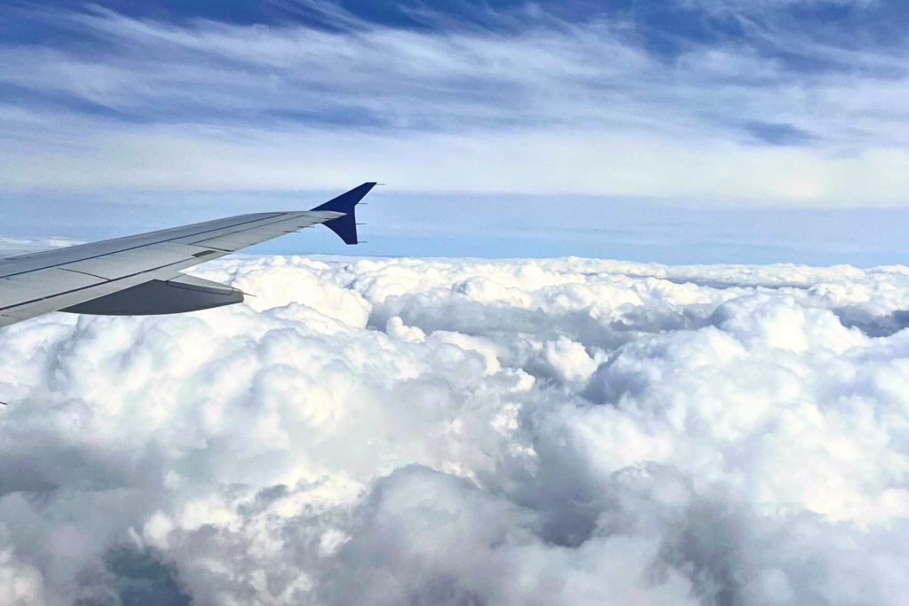 View from an airplane window showing a wing above a layer of fluffy white clouds with a blue sky and wispy clouds above.