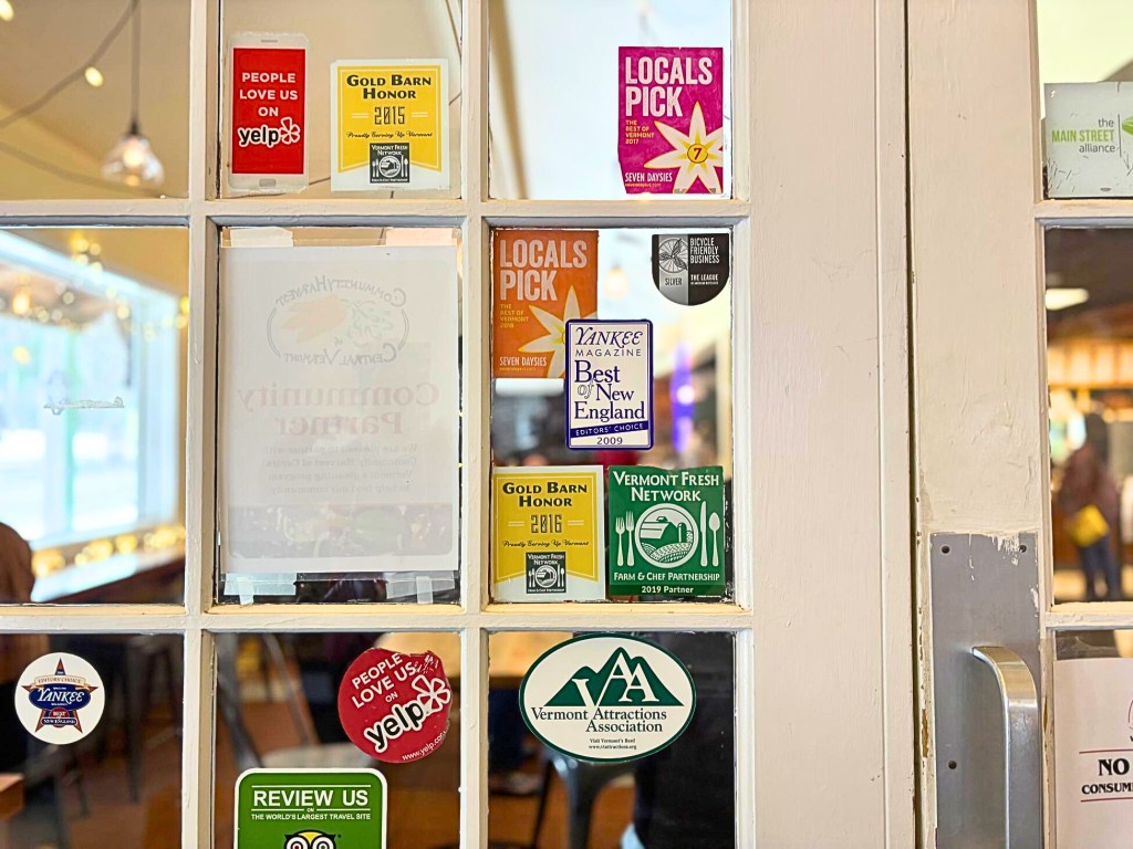 Glass door covered with various award, review, and association stickers, including Yelp, Vermont Fresh Network, and Yankee Magazine. Restaurant interior visible through the glass.