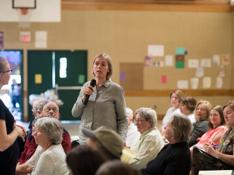 A woman stands and speaks into a microphone during a community meeting in a room with seated attendees and decorated walls.