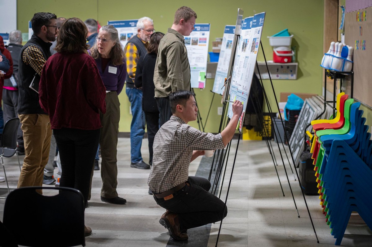 Several people examine and discuss posters on easels at an indoor event, while one person kneels to adjust a display. Colorful stacked chairs are visible on the right.