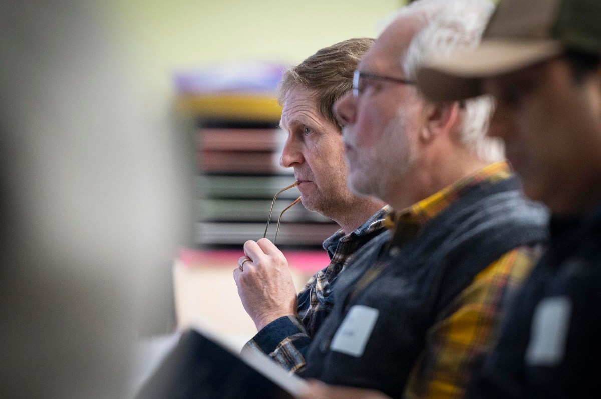Three men sit in a row, one holding his glasses to his mouth, appearing to listen attentively to something out of view.