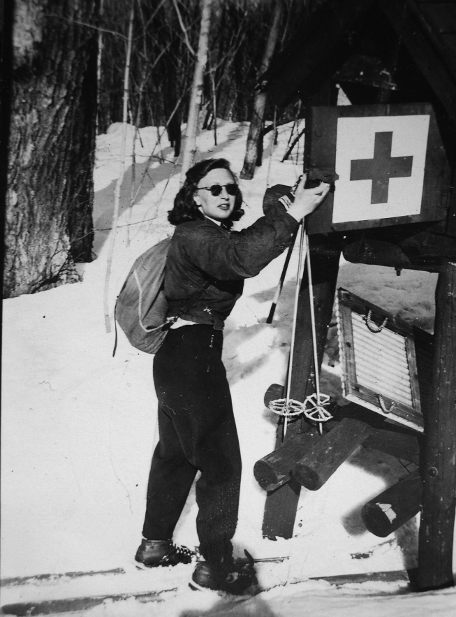 A woman in winter clothing and skis stands beside a first aid box with a red cross sign, adjusting or inspecting it in a snowy, wooded area.