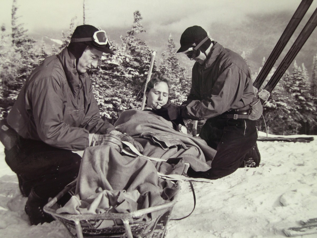 Two people in winter clothing attend to an injured person lying on a sled in a snowy outdoor setting with trees and mountains in the background.