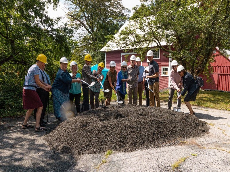 A group of people wearing hard hats and holding shovels stand around a pile of gravel outdoors, participating in a groundbreaking ceremony.