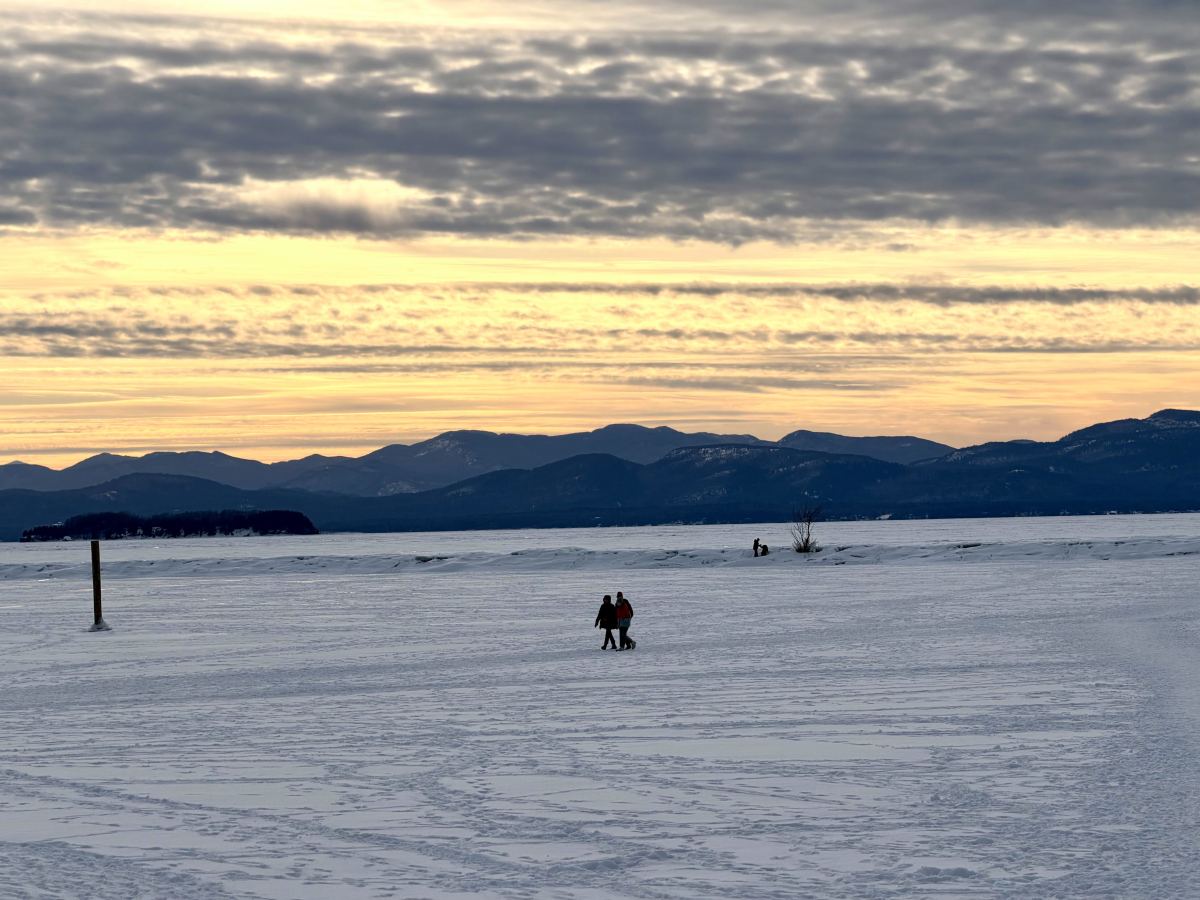 Two people walk across a snowy, frozen landscape with distant mountains and a cloudy sky at sunset.