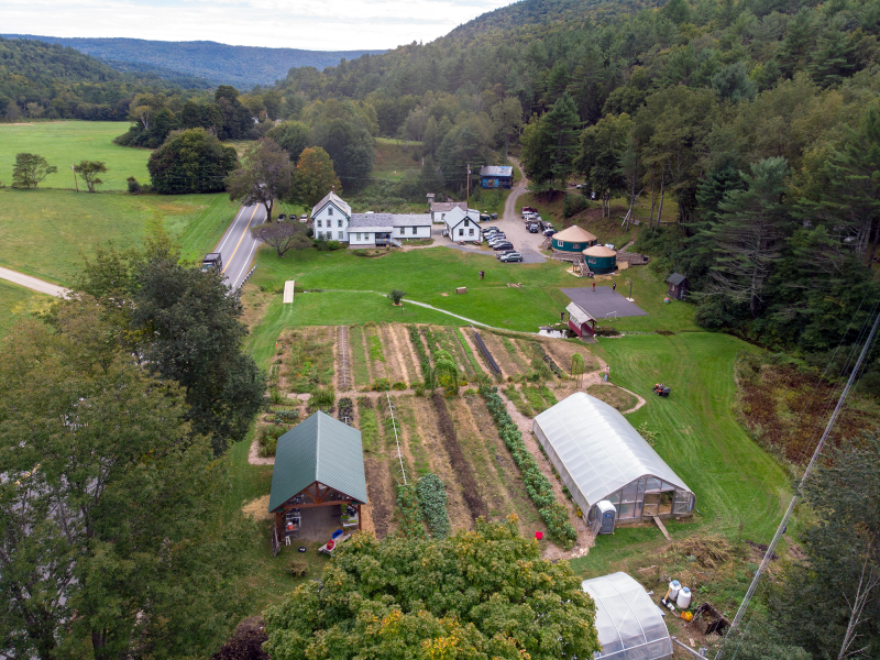 Aerial view of a rural farm with garden plots, greenhouses, several buildings, parked cars, and surrounding forested hills.