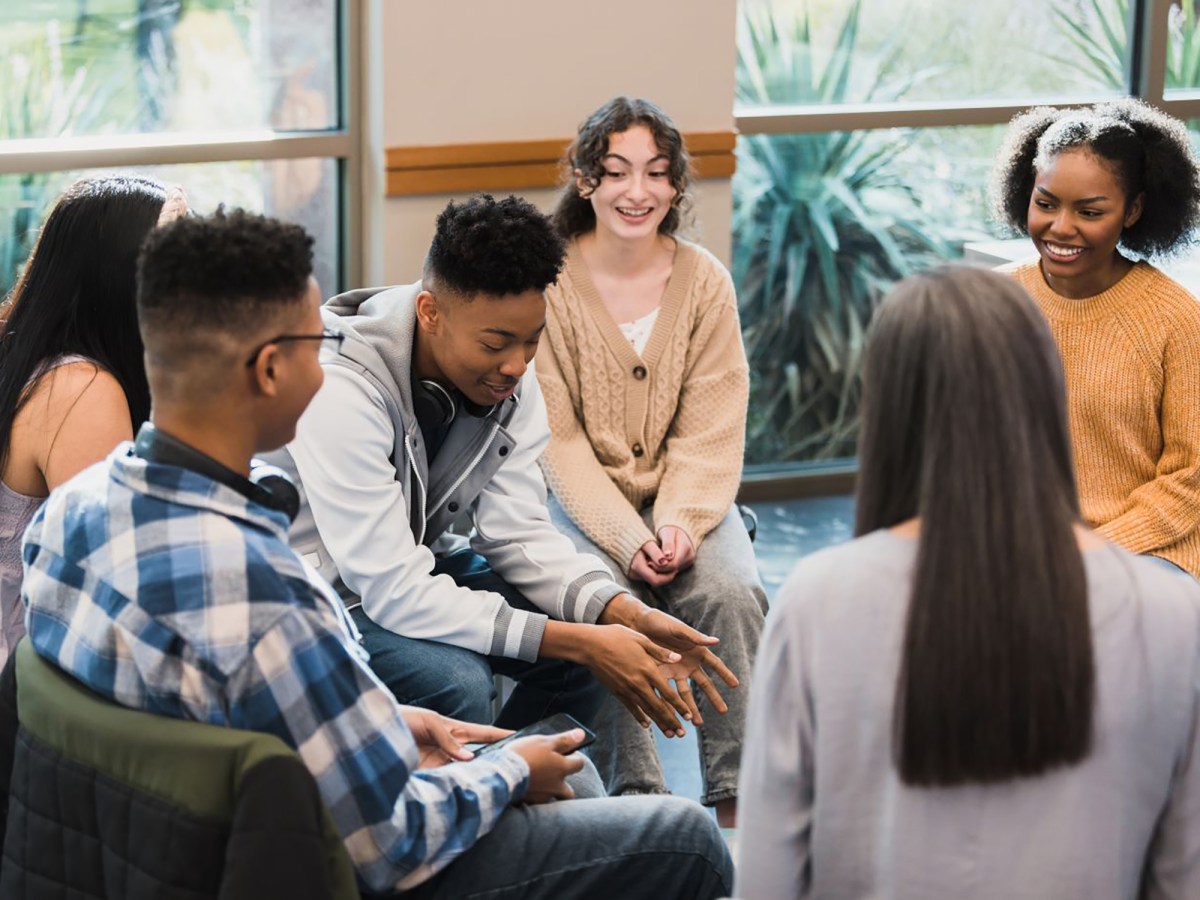 A group of six young adults sit in a circle indoors, engaged in conversation and smiling, with large windows in the background.