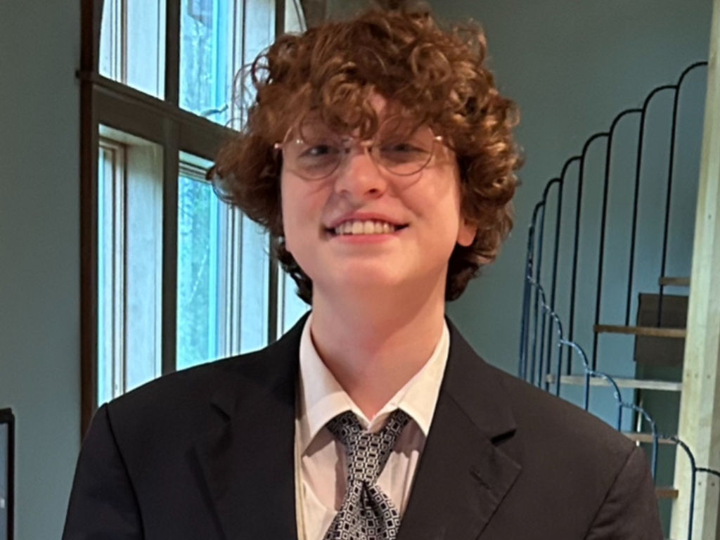 A person with curly hair, glasses, and a suit stands indoors near a window and a staircase, smiling at the camera.