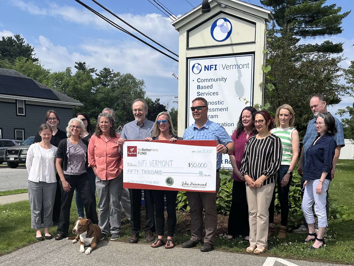 A group of people stand outside next to an NFI Vermont sign, holding a large check for $50,000 from Dealer.com/ Cox Automotive; a dog sits at their feet.