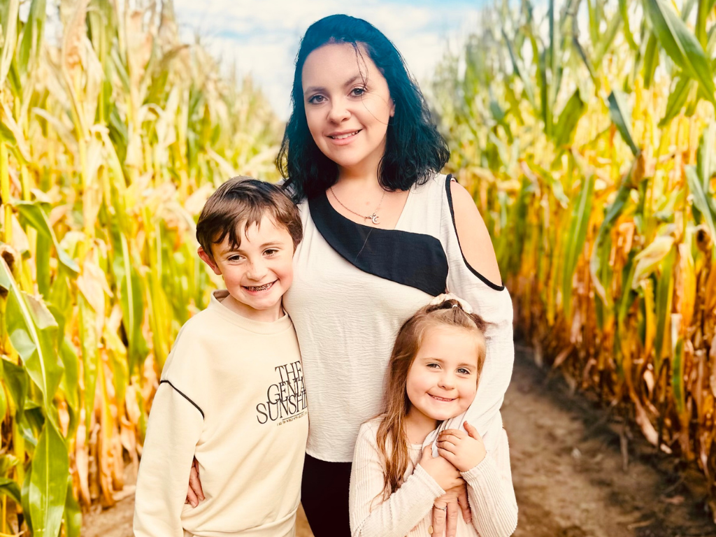 A woman stands in a cornfield with two smiling children, one boy and one girl, on a sunny day.