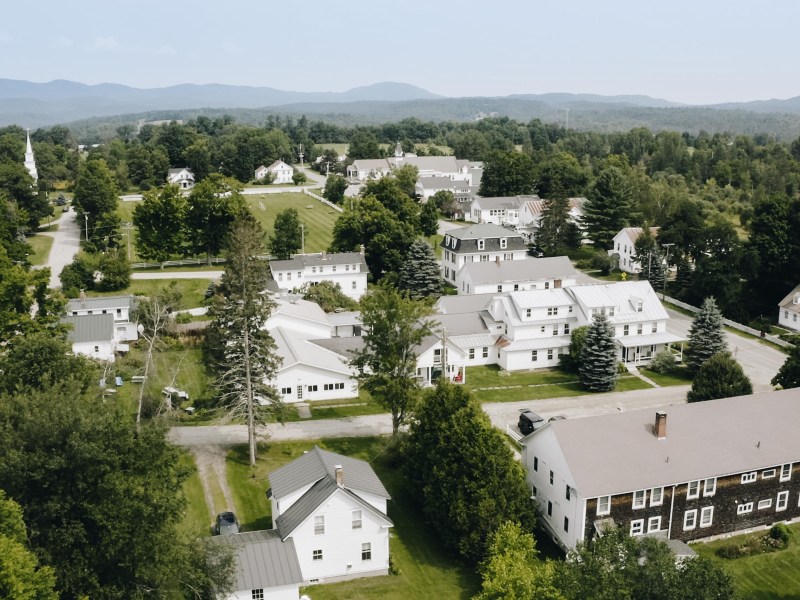 Aerial view of a small rural town with white houses, green trees, and distant hills under a clear sky.