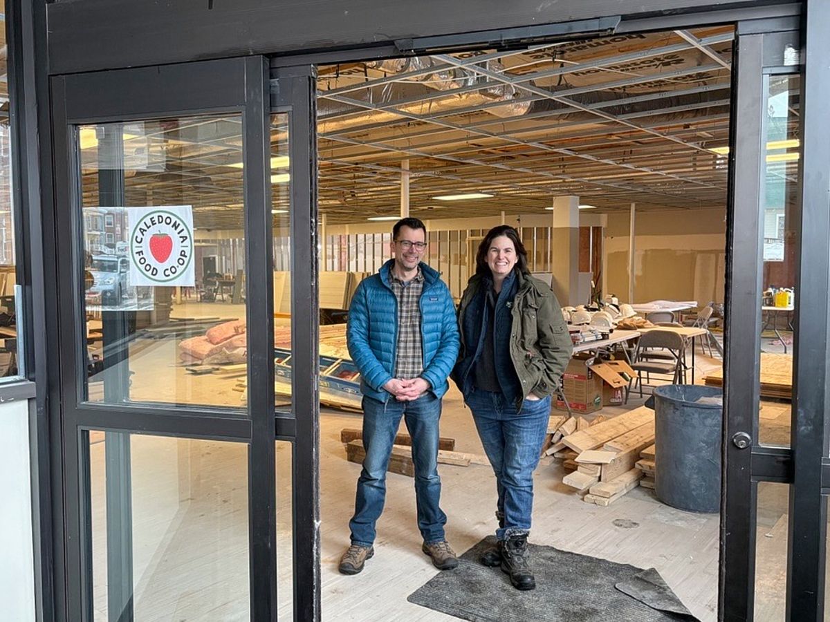 Two people stand inside an unfinished commercial space with construction materials, tools, and tables visible. A "Co-op" sticker is on the glass door.