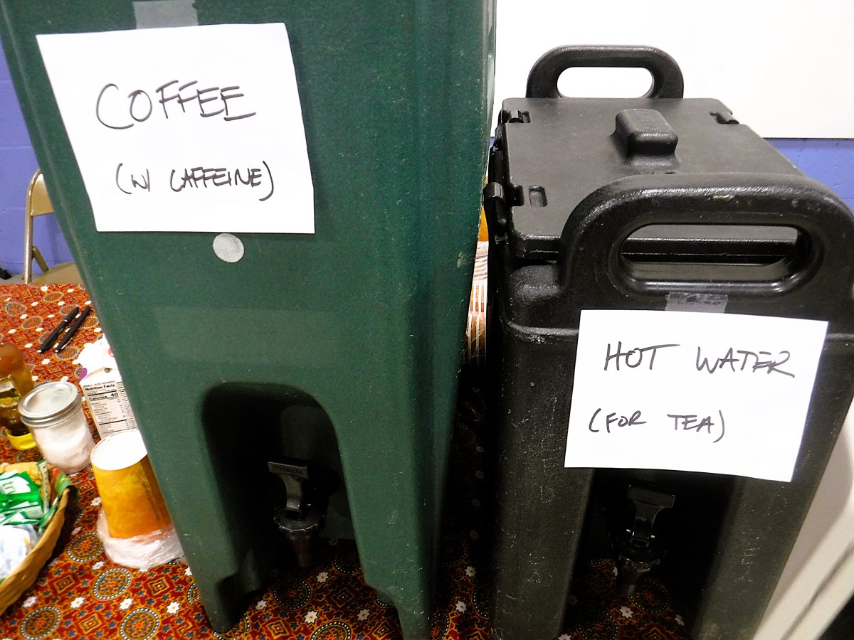 Two beverage dispensers labeled "Coffee (w/ caffeine)" and "Hot Water (for tea)" sit on a table with cups, tea bags, and condiment packets.