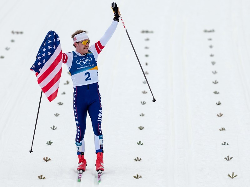 A cross-country skier wearing a number 2 bib holds an American flag and raises an arm while skiing on snow, with Olympic rings visible on the uniform.