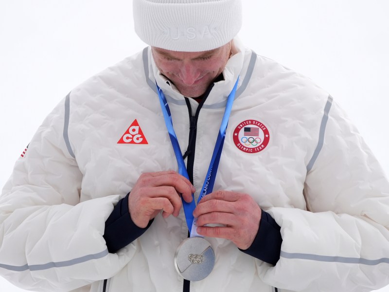 An athlete in a white Team USA jacket and beanie looks down while holding a silver Olympic medal on a blue ribbon around their neck.