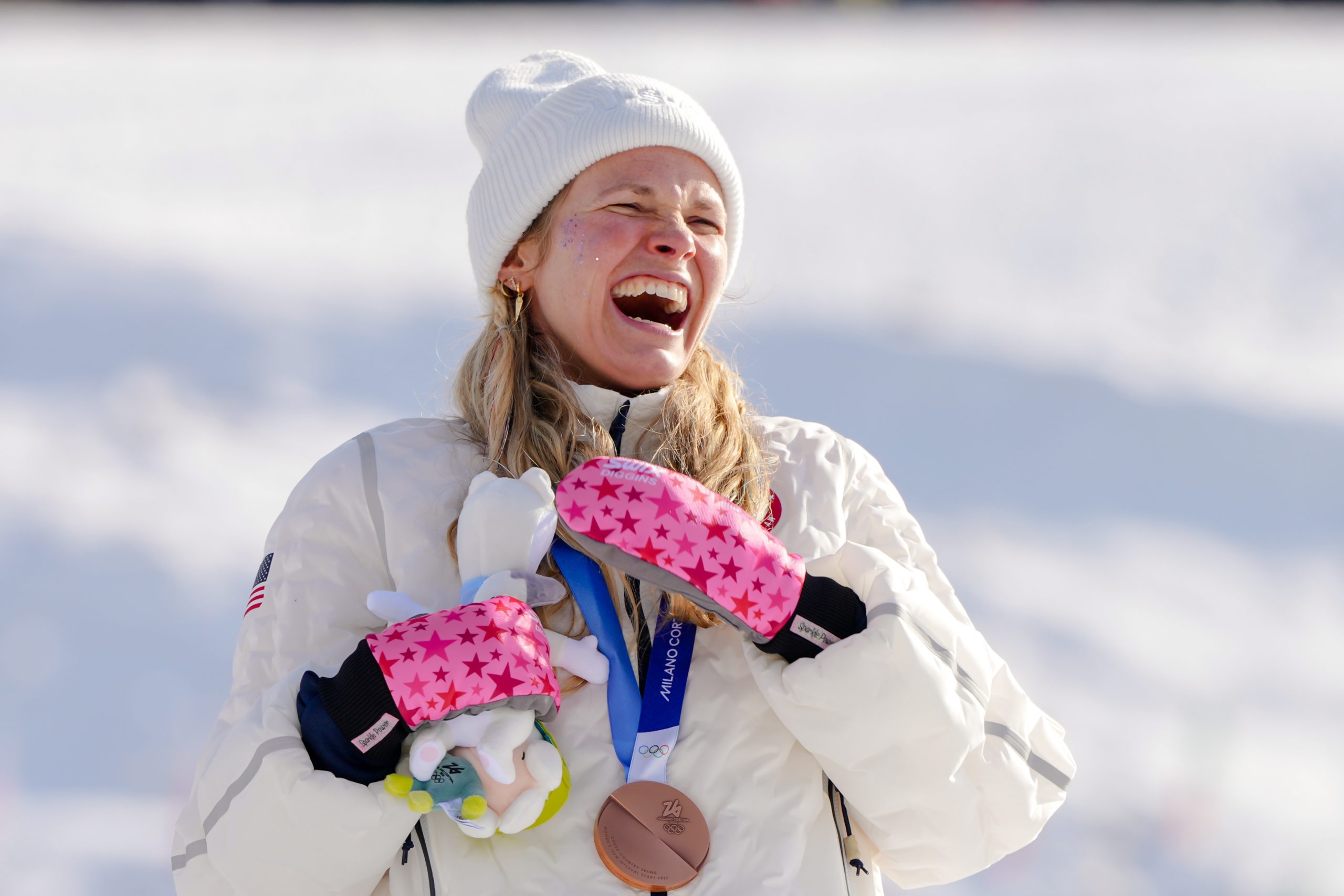A person in winter clothing and star-patterned mittens smiles while holding a mascot and wearing a bronze medal on a snowy background.