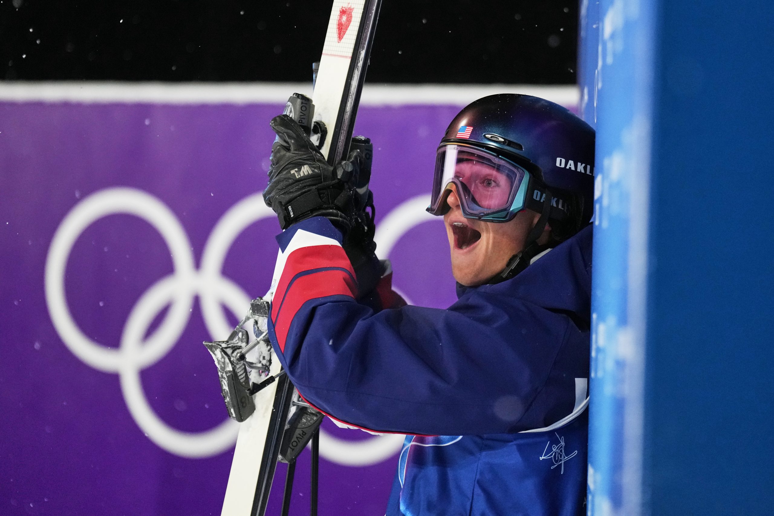 A skier in blue winter gear and a helmet holds skis and looks surprised or excited near an Olympic symbol backdrop.