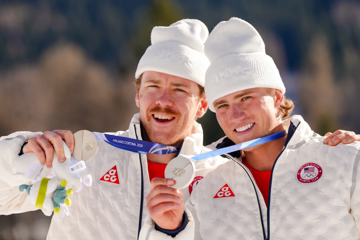 Two athletes in white jackets and hats smile and display silver medals at the 2022 Winter Olympics, with a blurred outdoor background.