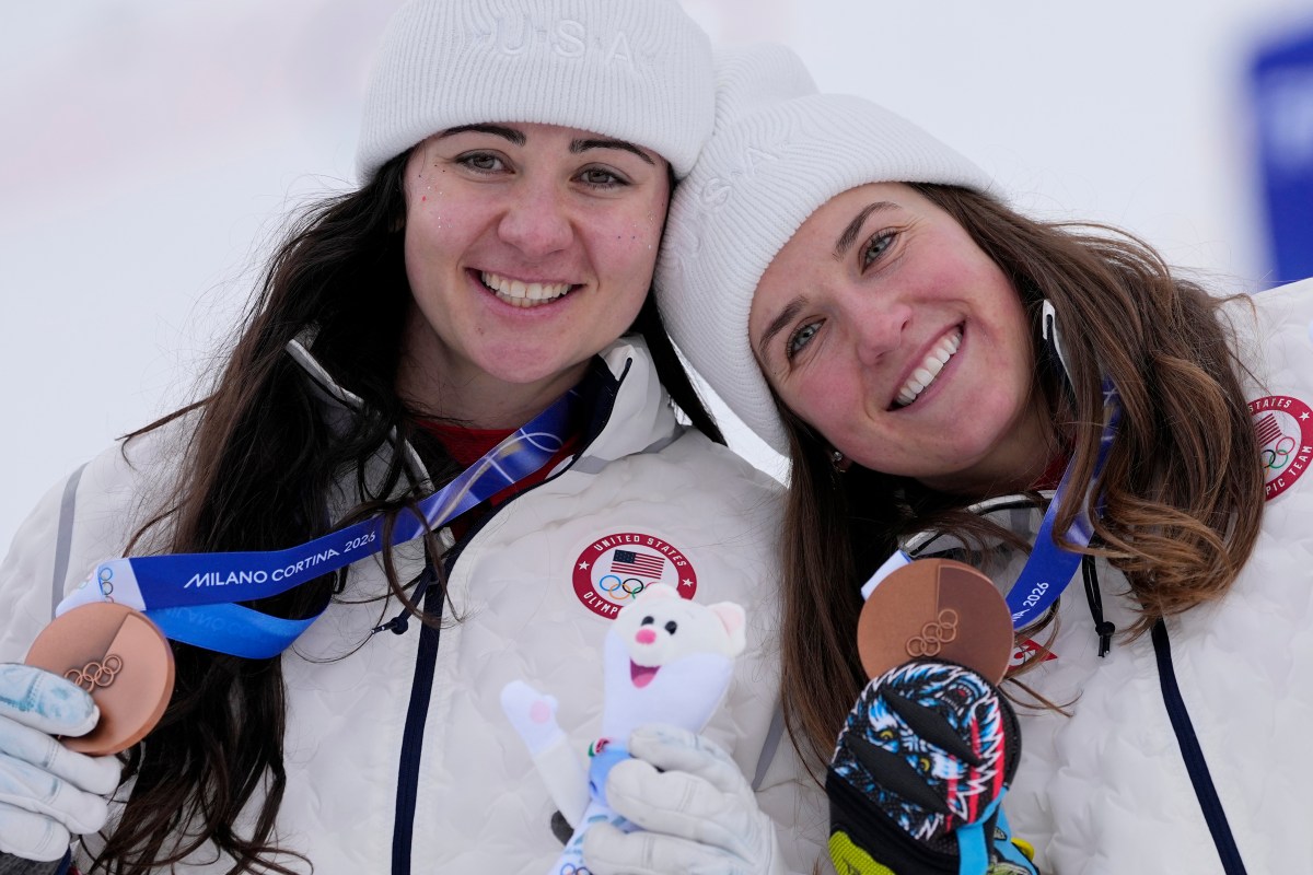 Two female athletes in white USA jackets and hats smile while holding bronze medals and a plush toy at a snowy event.