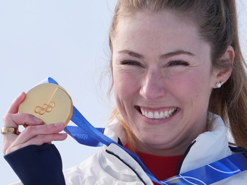 A woman smiling and holding up a gold Olympic medal on a blue ribbon.
