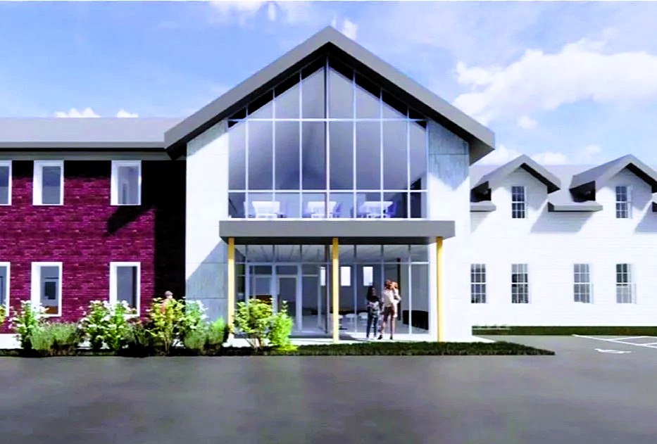 Two-story modern building with large glass entrance, pitched roof, and a mix of red brick and white siding; a person stands near the main doors.